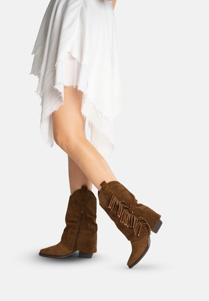 Gambe in una gonna bianca asimmetrica che indossano stivali marroni in suede a metà polpaccio con frange e dettagli con cerniera su sfondo bianco.