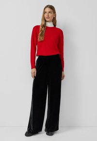 Rode sweater met lange mouwen, gecombineerd met zwarte fluwelen wijde broek en zwarte schoenen, tegen een eenvoudige achtergrond.