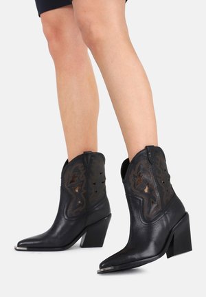 Nogi w czarnych skórzanych kowbojskich butach z ozdobnymi wycięciami, spiczastymi czubkami, blokowymi obcasami i metalowymi nasadkami na palcach na białym tle.