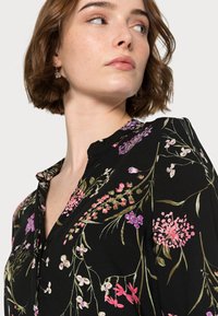 Blusa preta de flores com mangas curtas, apresentando flores bordadas em rosa e roxo, colarinho mandarina e detalhe de botões na frente.