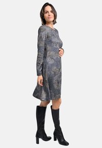 Graues langärmeliges Kleid mit einem ausgestellten Rock und einem Blättermuster. Kombiniert mit knielangen schwarzen Stiefeln mit Absatz. Glattes, strukturiertes Material.