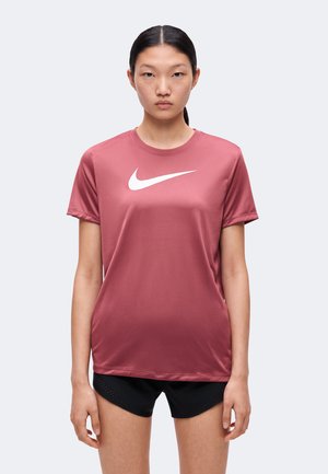 Jeune femme aux cheveux raides porte un t-shirt de sport bordeaux à manches courtes avec un logo Nike blanc et un short de sport noir, face à nous.