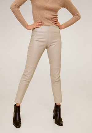 Osoba ubrana w beżowe, skórzane spodnie z wysokim stanem, zestawione z beżowym swetrem z warkoczem i czarnymi, wysokimi botkami na obcasie, stojąca z rękami na biodrach.