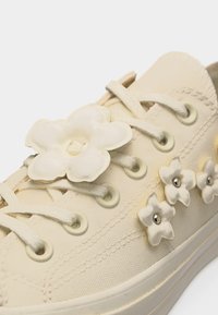 Μπεζ πάνινο αθλητικό παπούτσι με λευκά κορδόνια και διακοσμητικές λευκές ανθοειδείς λεπτομέρειες κατά μήκος της πλευράς και στο πάνω μέρος κοντά στα οπές των κορδονιών.