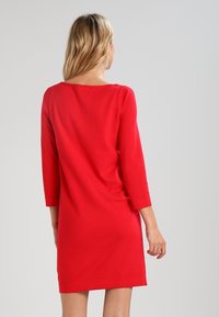 Robe rouge, longueur genou avec des manches trois-quarts. Tissu lisse et extensible. Encolure ronde simple, sans embellissements notables.