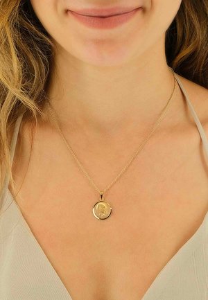 Collier en pendentif en or avec un charme circulaire présentant un design de profil, suspendu à une chaîne délicate, porté sur un haut de couleur claire.
