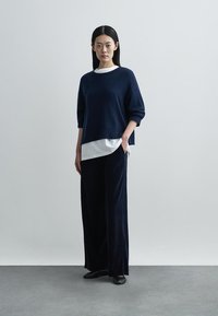 Oversized marineblauwe trui over een wit shirt, gecombineerd met wijde, donkere broek en zwarte platte schoenen, tegen een neutrale achtergrond.