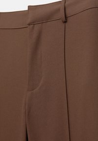 Gros plan d’un pantalon marron sur mesure montrant la taille, la passants de ceinture, la couture et les détails des poches sur un tissu lisse.