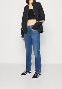 Chaqueta negra con cremallera y logo blanco, top corto negro, jeans azules de tiro medio con cremalleras laterales y zapatillas negras con rayas blancas.
