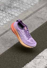 Zapatilla de correr morada con parte superior de malla, entresuela naranja contrastante, cordones planos y suela texturizada, mostrando un diseño en capas y detalles reflectantes.