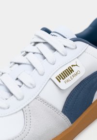 Baskets Puma Palermo blanches avec lacets blancs, bande en daim bleu, accents en daim gris et semelle en gomme, affichant un logo Puma doré sur le côté.