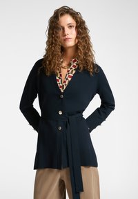 Cárdigan de punto azul marino con un cinturón en la cintura, dos botones frontales y dobladillos acanalados, superpuesto a una blusa estampada con formas geométricas en rojo y beige.