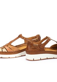 Sandalias de cuero marrón con costuras decorativas, laterales perforados y plantilla color beige. Presentan una suela de cuña blanca y detalles metálicos.