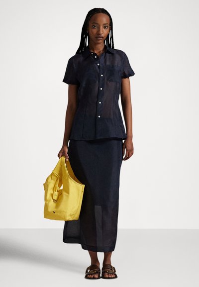 Sieviete tērpusies caurspīdīgā tumši zilā pogājamas krekla un atbilstoša svārku komplektā, tur rokās dzeltenu somiņu, valkā brūnas sandales, stāv uz vienkārša fona.