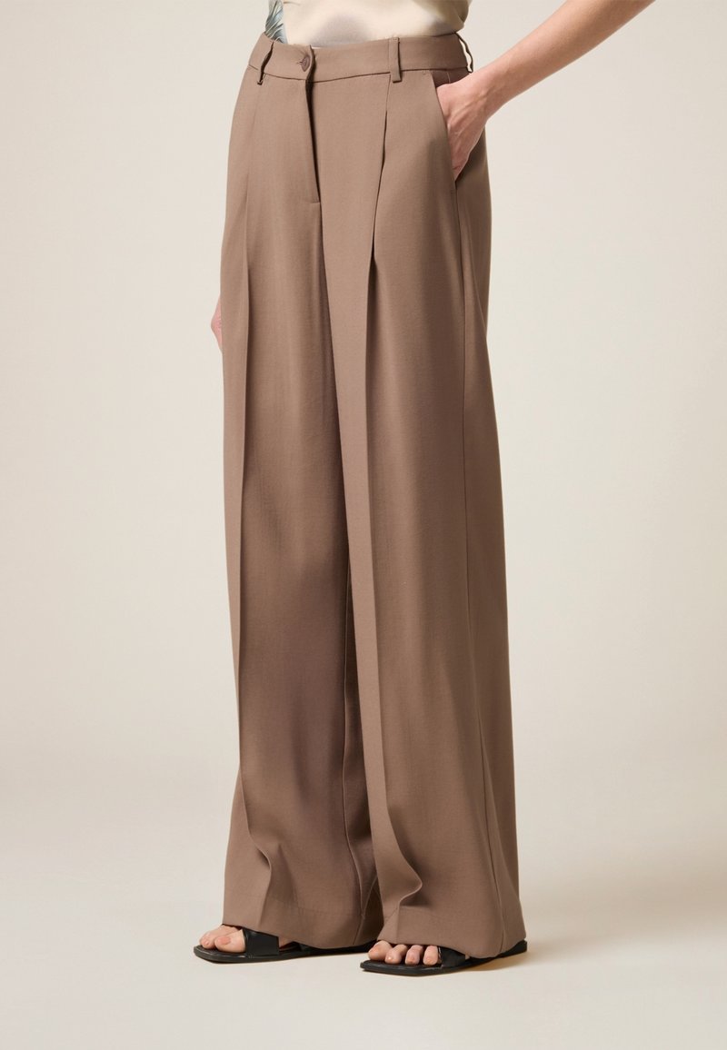 Weit geschnittene Hose aus einem hellbraunen Stoff, mit Falten, hoher Taille und seitlichen Taschen. Kombiniert mit flachen schwarzen Sandalen.
