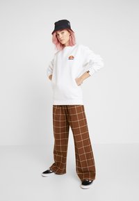 Sudadera blanca con un logo circular, pantalones de pierna ancha a cuadros marrones y un sombrero tipo cubo negro. Zapatillas negras con acentos en la suela blanca.