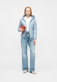 Γυναίκα που φοράει ανοιχτό μπλε φουσκωτό μπουφάν, λευκό πουκάμισο, τζιν παντελόνι με φαρδιά σκισμένα μπατζάκια, κρατώντας μια μικρή κόκκινη τσάντα, στέκεται σε λευκό φόντο.