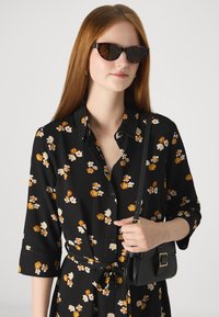 Mustree lilleline kleit kollaste ja valgete lilledega, nööpidega esiküljel, pikad varrukad, seotud vöökoht, koos väikese musta käsipagasiga ja prillidega.