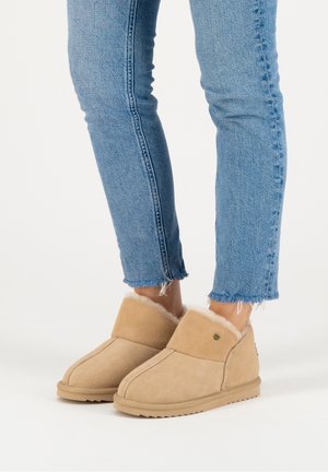 Personne portant un jean bleu clair et des bottes d’hiver beige doublées de fourrure, debout sur une surface blanche.