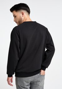 Felpa nera con collo rotondo e maniche raglan, presenta polsini e orlo a coste. Texture liscia, indossata con jeans grigi.