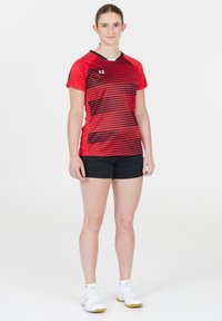 Röd och svart randig atletisk tröja med korta ärmar, i kombination med svarta shorts. Modellen bär vita sportskor. Enfärgad bakgrund.