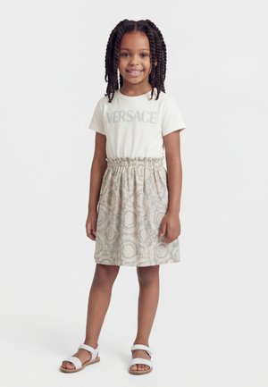 Giovane ragazza in piedi e sorridente, che indossa una t-shirt bianca Versace, gonna a fantasia e sandali bianchi su sfondo neutro.