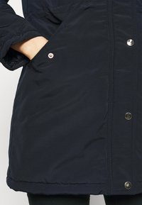 Navy blue parka with a smooth fabric texture, featuring side pockets, silver snap buttons, and a slightly longer hemline.