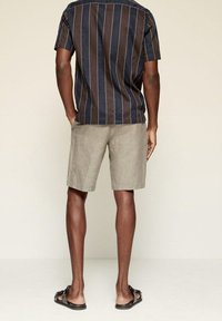 Camisa de manga corta con rayas verticales azules y marrones, combinada con pantalones cortos gris claro y sandalias negras, vista desde atrás.