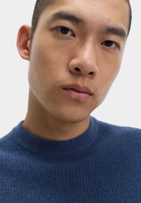 Modrý pletený svetr s texturovaným povrchem a žebrovaným výstřihem, který nosí člověk s krátkými vlasy a neutrálním výrazem.