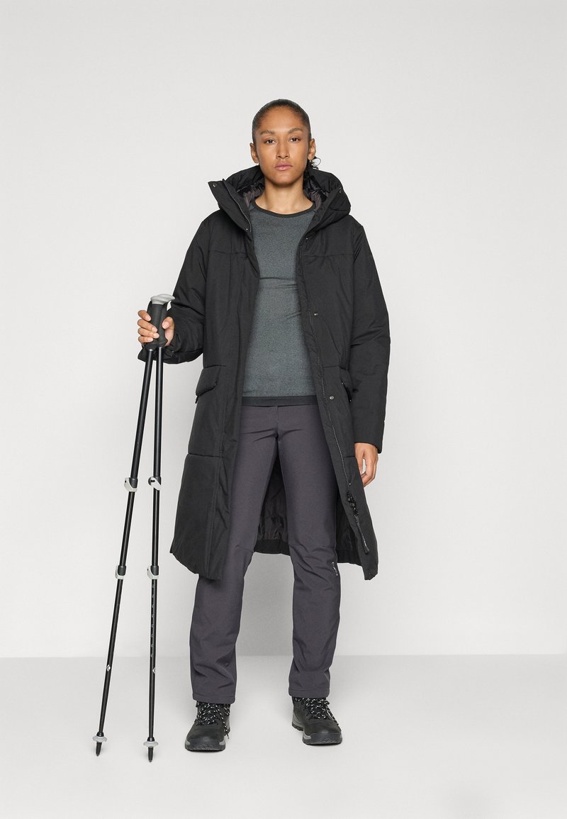 Frau in einem langen schwarzen Mantel, grauem Pullover, dunkler Hose und Wanderschuhen, die zwei schwarze Trekkingstöcke vor einem weißen Hintergrund hält.