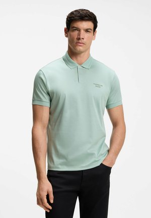 Homme portant un polo à manches courtes vert clair avec un petit logo sur la poitrine et un pantalon noir, debout avec une main dans la poche.
