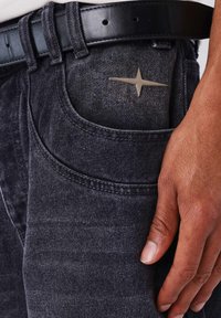 Vaqueros de mezclilla negros con un acabado texturizado, que presentan un prominente adorno en forma de estrella plateada en el bolsillo trasero y un cinturón de cuero negro.