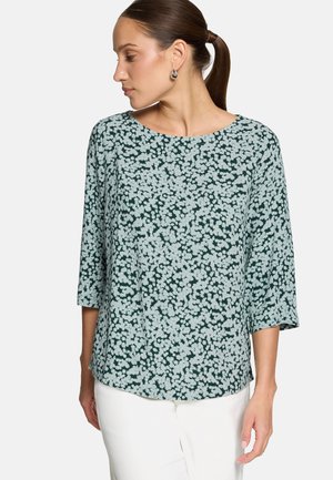 Femme aux cheveux bruns attachés, portant des boucles d'oreilles argentées en anneau, un chemisier à manches longues à fleurs vert foncé et bleu clair, et un pantalon blanc, regardant vers la gauche.