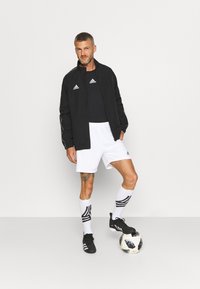 Giacca sportiva nera con zip, pantaloncini bianchi, scarpini da calcio neri, calze bianche con strisce nere e un pallone da calcio su uno sfondo bianco.