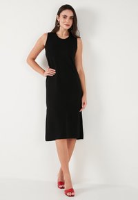 Black sleeveless dress with a fitted silhouette, knee-length hem, smooth texture, paired with red open-toe heels and simple jewelry.