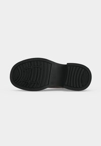 Semelle en caoutchouc noire d'une chaussure avec un motif de la bande de roulement pour l'adhérence, montrant clairement les zones du talon et de l'avant sur un fond uni.