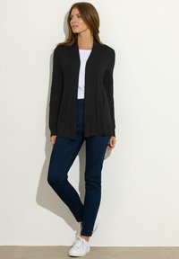 Czarny długi kardigan z odkrytym przodem, o prążkowanej fakturze, zestawiony z ciemnoniebieskimi dopasowanymi dżinsami i białymi trampkami. Prosty, casualowy zestaw.