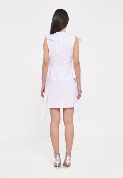 Robe blanche sans manches au design ajusté, dotée de liens latéraux et d'une fermeture éclair dans le dos. Le mannequin porte des sandales à talons hauts métalliques.