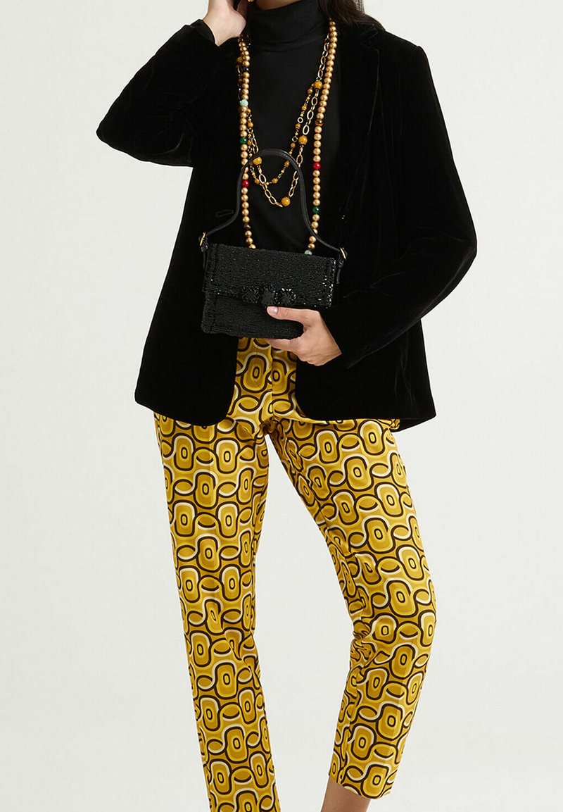 Blazer en velours noir porté sur un col roulé noir, associé à un pantalon jaune à motifs géométriques et un sac à main noir texturé.