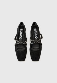 Chaussures noires à bout carré avec texture tissée, dotées de sangles croisées ornées d'accents métalliques circulaires. Logo de la marque visible à l'intérieur.