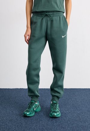 Person trägt dunkelgrüne Nike Jogginghose und passende grüne Nike Sneaker und steht auf blauem Teppich.