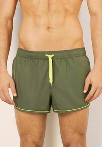 Olivgröna badshorts med elastisk midja och neongul snodd. Har kontrasterande gult kantband längs fållen och sidorna.