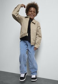 Beige ritsjack met een cropped ontwerp, een zwart t-shirt eronder, lichtblauwe corduroy broek en zwarte sneakers met witte veters.