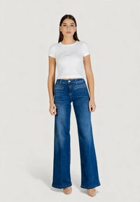 Valkoinen ribbineulottu lyhythihainen paita, jossa on "Guess"-logo, yhdistettynä sinisiin leveälahkeisiin farkkuihin. Sileä pinta, normaali vyötärönauha, farkuissa kevyt haalistuma.