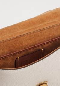 Int érieur d'un sac à main en cuir blanc avec un doux revêtement en daim tan, comprenant une poche pour cartes bordeaux et un accent en métal doré sur le bord.