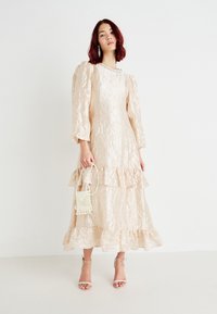 Krøllad beige kjole med puffede ærmer og volangkant, parret med en lille hvid vævet taske og strappy beige hæle.