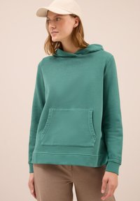 Joven con una sudadera teal con bolsillo frontal y una gorra beige, posando frente a un fondo claro y liso.