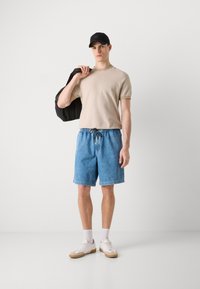 T-shirt beige clair à manches courtes, shorts en denim bleus, casquette noire, chaussures blanches avec semelles en caoutchouc, et sac noir porté sur l'épaule.