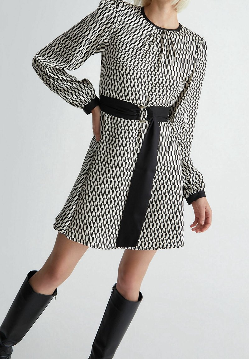 Robe à motifs noir et blanc avec une taille cintrée, des manches longues bouffantes et une ceinture noire contrastante. Tissu lisse avec une finition soignée.