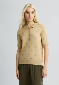 Tan korte mouwen polo trui met een gestructureerd logo patroon, drie knopen en een geribbelde kraag. Gecombineerd met olijfgroene broek.